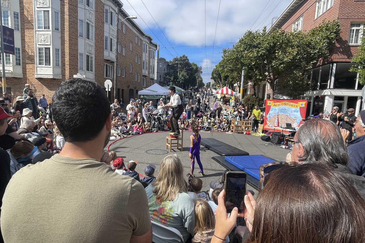 Street performance with a crowd watching a balancing act in a sunny urban setting.