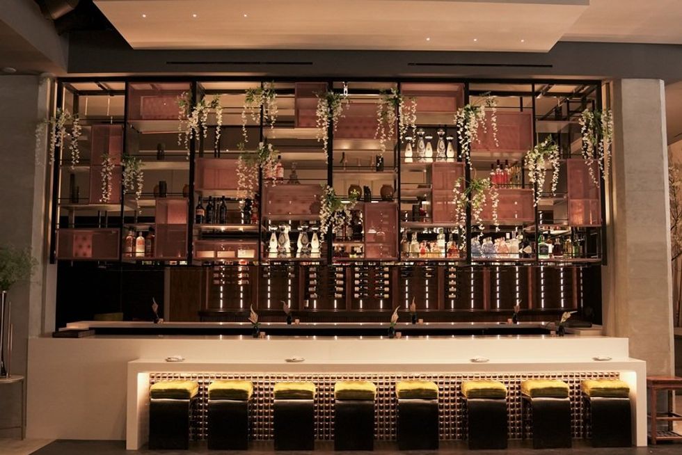 Stylish bar interior with hanging plants, backlit shelves, and green cushioned stools.