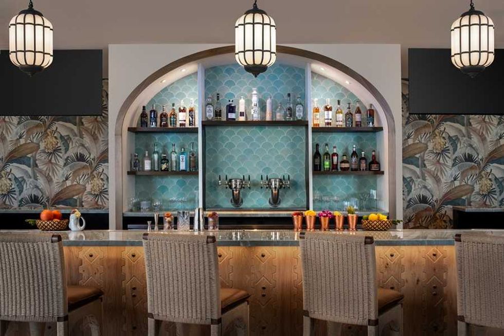 Stylish bar with wicker chairs, blue tile, bottles, flowers, and fruits under hanging lamps.