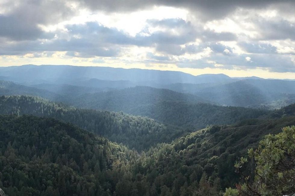 Sun rays breaking through clouds over a vast, forested mountain landscape.