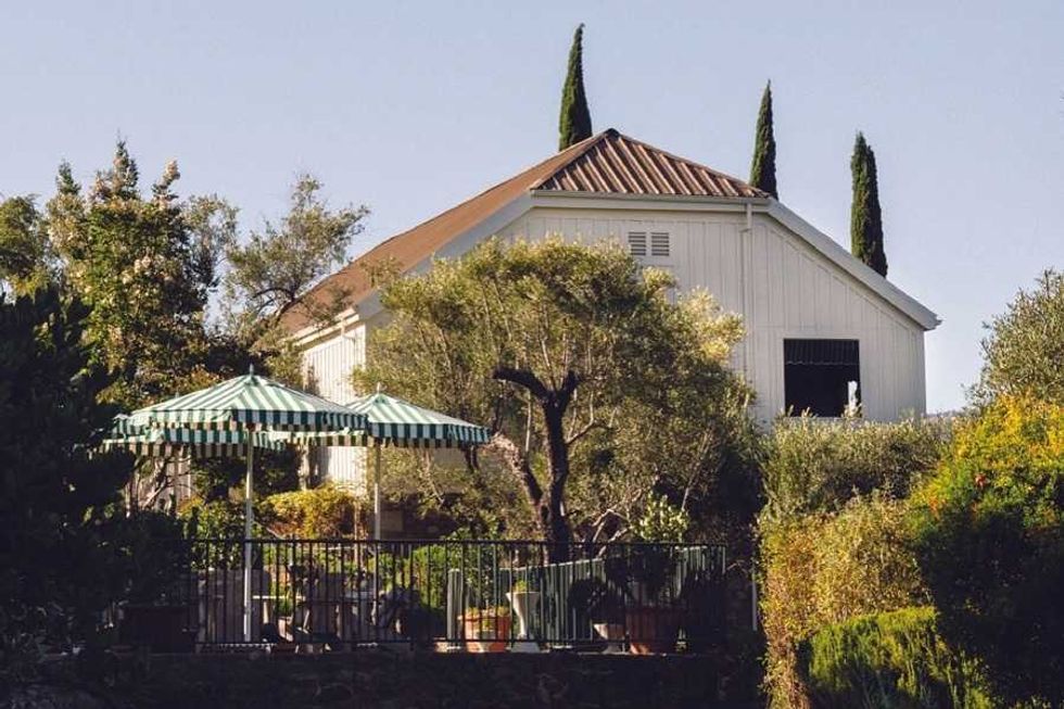 Sunny patio with striped umbrella, chairs, and lush greenery outside a white barn-style building.