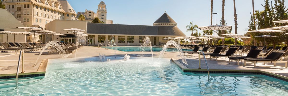 Sunny poolside with loungers, umbrellas, fountains, and palm trees beside a grand building.