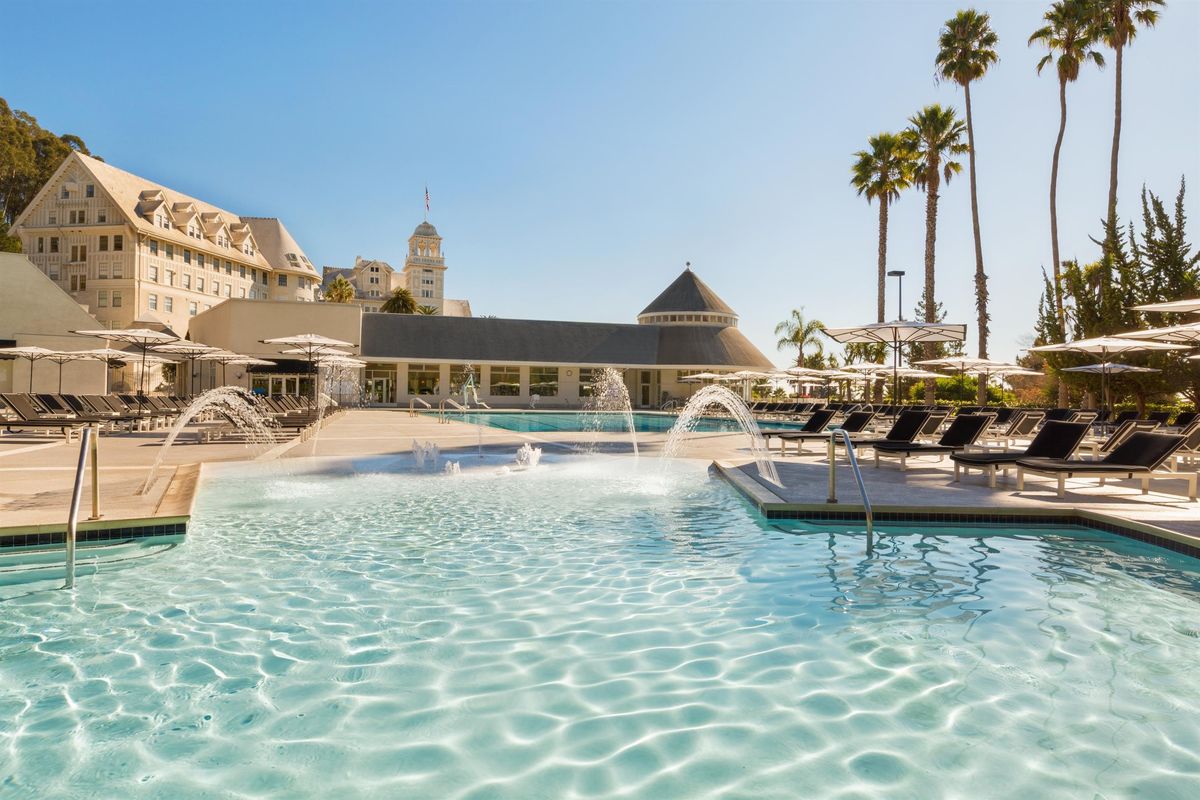 Sunny poolside with loungers, umbrellas, fountains, and palm trees beside a grand building.