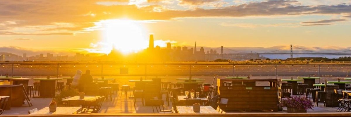 Sunset over city skyline with clouds, viewed from an outdoor patio with tables and chairs.