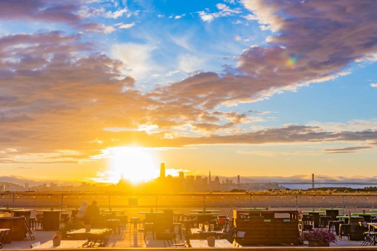 Sunset over city skyline with clouds, viewed from an outdoor patio with tables and chairs.