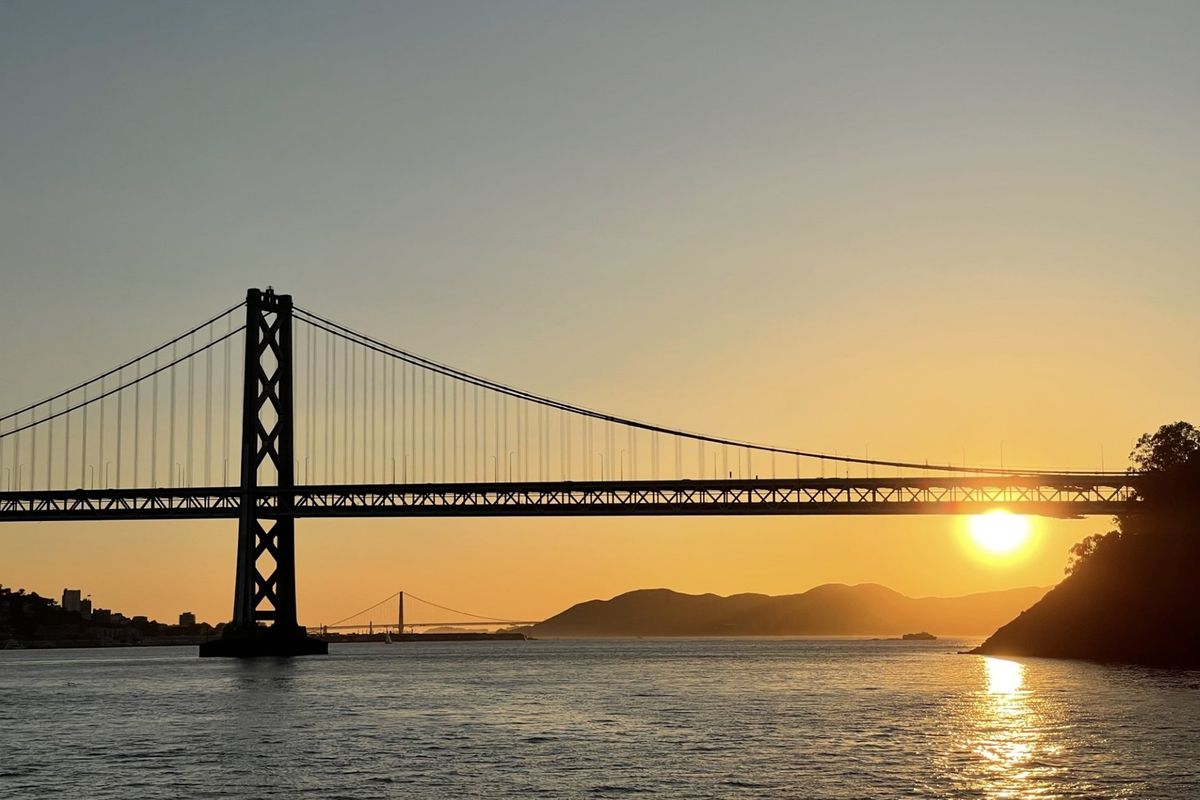 Sunset view of a suspension bridge over calm water with silhouetted hills in the background.