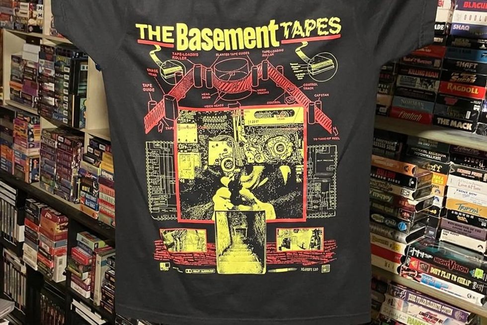 T-shirt with "The Basement Tapes" graphic, surrounded by VHS tapes on shelves.
