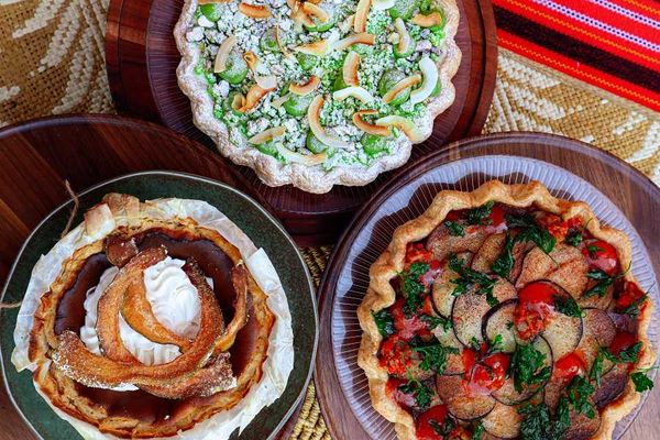 Three assorted savory pies on wooden plates, topped with vegetables and cream.