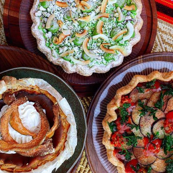 Three assorted savory pies on wooden plates, topped with vegetables and cream.