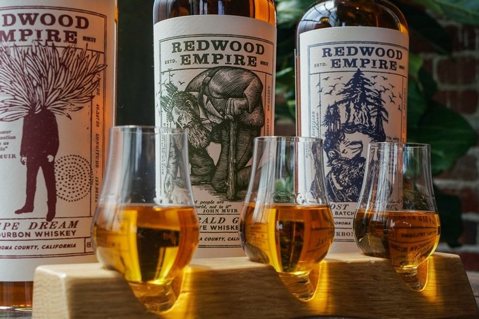 Three glasses of whiskey in front of Redwood Empire whiskey bottles with artistic labels.
