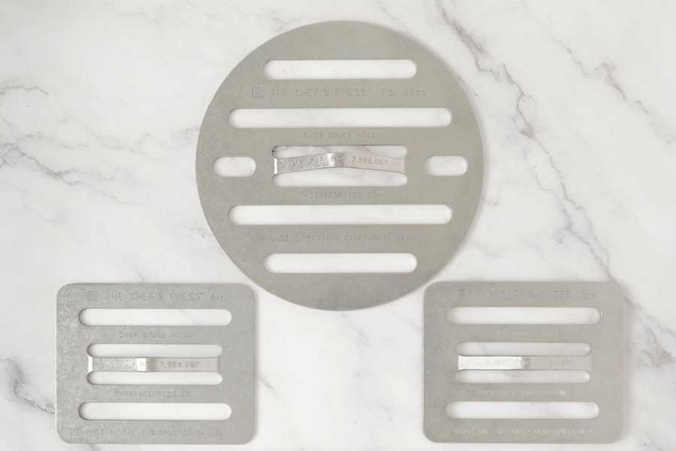 Three metal grill weights on a marble surface, two rectangular and one round.