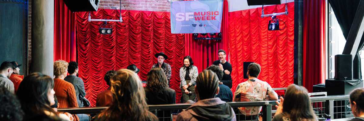 three people on stage at an SF Music Week event with a red curtain behind them and an audience
