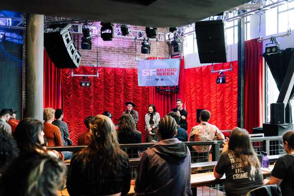 three people on stage at an SF Music Week event with a red curtain behind them and an audience