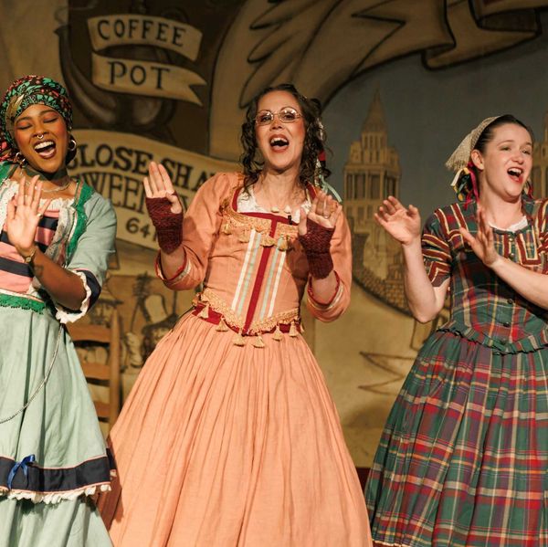 Three women in colorful historical costumes singing joyfully on stage.