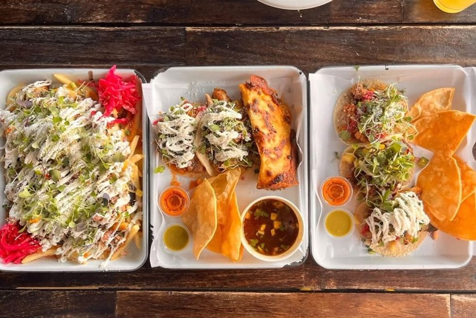 Trays with loaded fries, tacos, nachos, and various dips on a wooden table.
