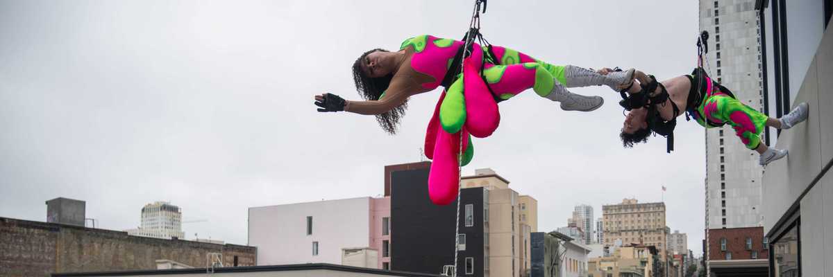 two acrobats in green and pink clothing hanging from the side of a tall building