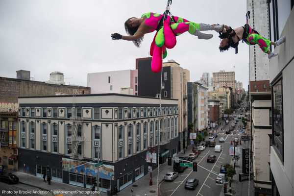 two acrobats in green and pink clothing hanging from the side of a tall building