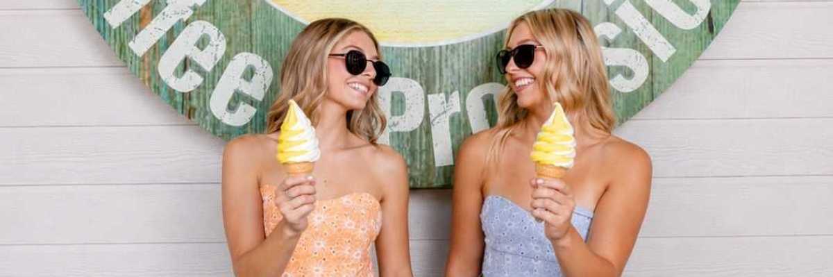 two blond girls in crop tops and sunglasses holding Dole Whip and looking at each other in front of a sign