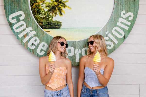 two blond girls in crop tops and sunglasses holding Dole Whip and looking at each other in front of a sign