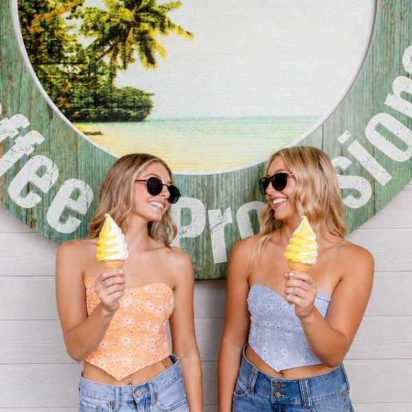 two blond girls in crop tops and sunglasses holding Dole Whip and looking at each other in front of a sign