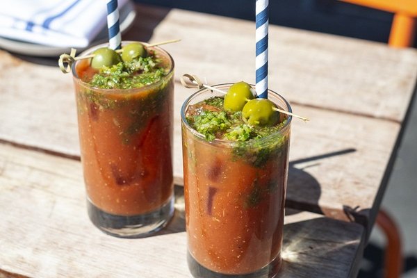 Two Bloody Mary cocktails with olives on a wooden table in the sunlight.
