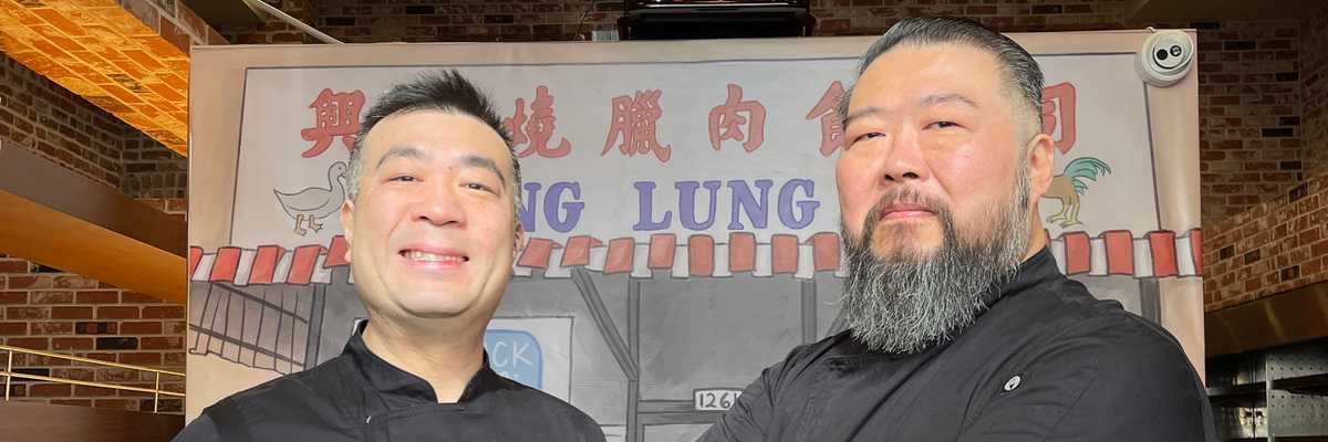 two Chinese American men smiling in front of a handpainted sign