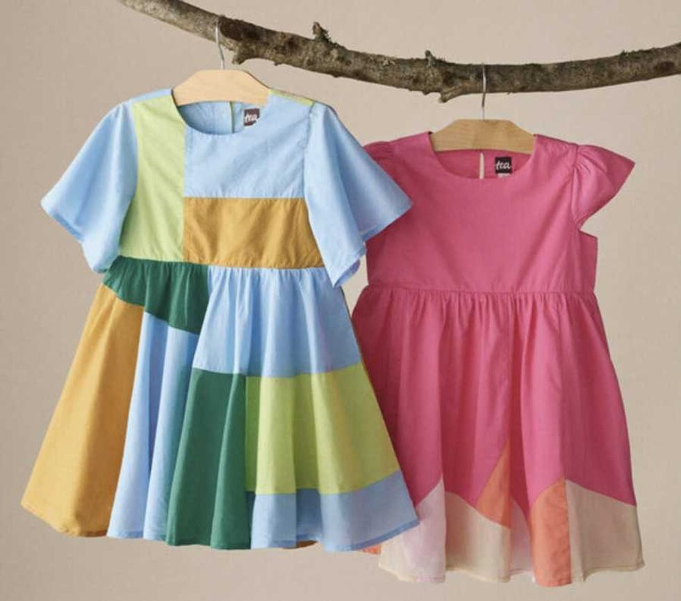 Two colorful children's dresses hang on a branch, one patchwork, one pink.
