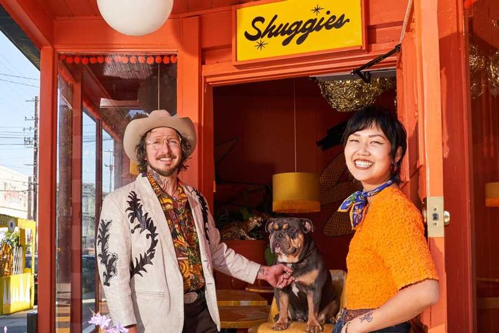 Two people and a dog in front of a brightly colored store named 