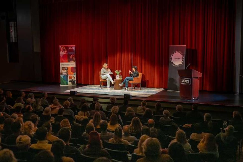 Two people having a discussion on stage with an audience and red curtains.