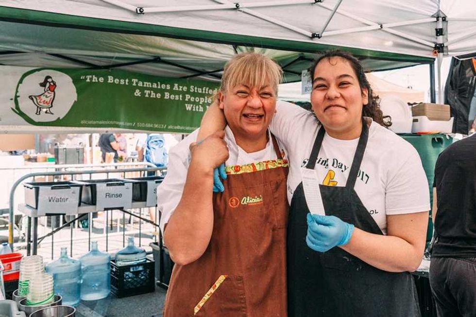 Two smiling women in aprons at a tamale stand under a canopy.