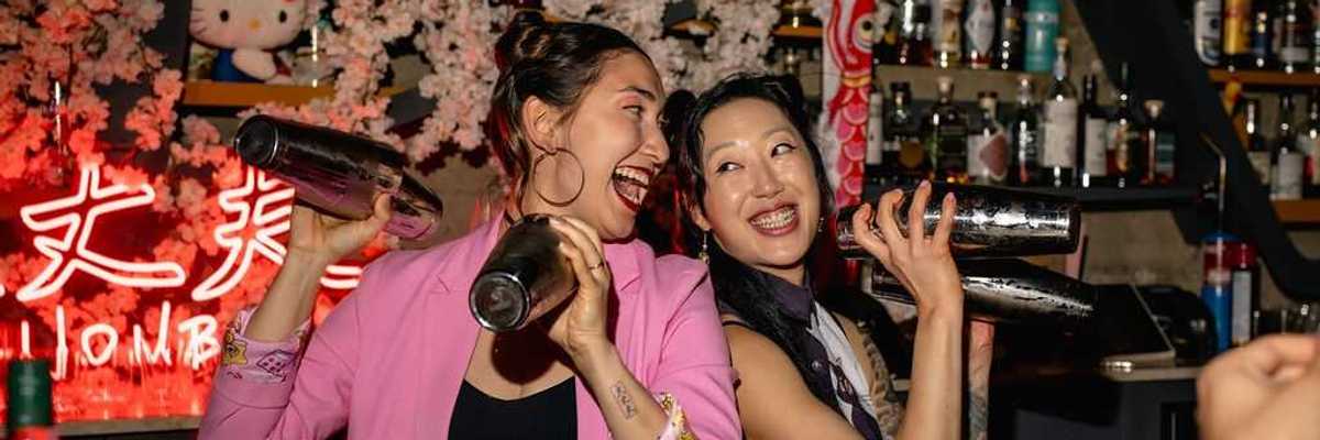 two women bartenders back to back smiling and shaking cocktail shakers