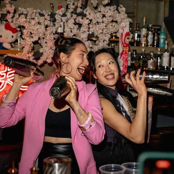 two women bartenders back to back smiling and shaking cocktail shakers