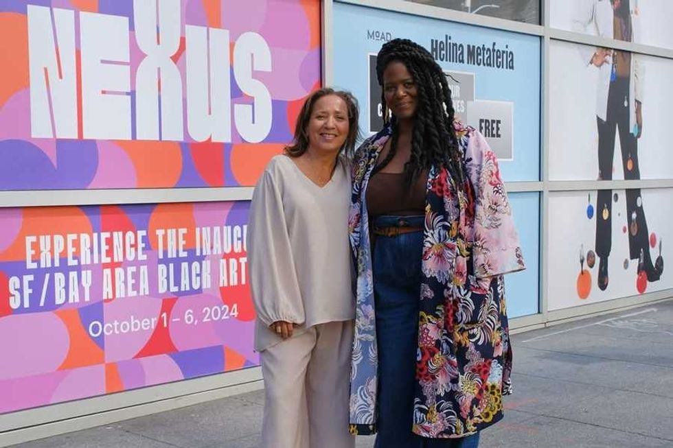 Two women smiling in front of a colorful "NEXUS" art event poster.