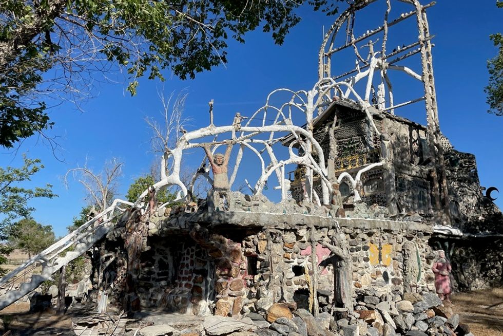 Unique stone and cement sculpture house with intricate white framework under a clear blue sky.