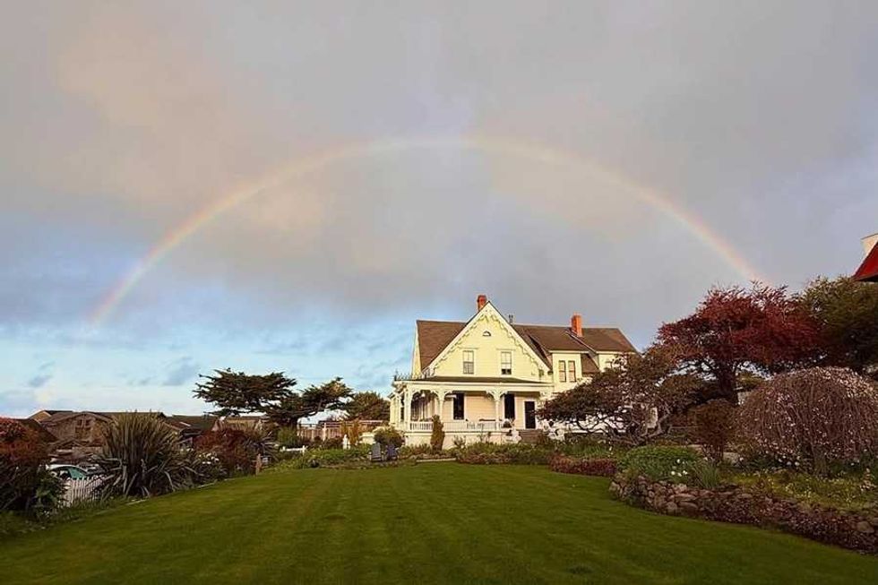 Victorian house under a rainbow with a lush green lawn and cloudy sky.