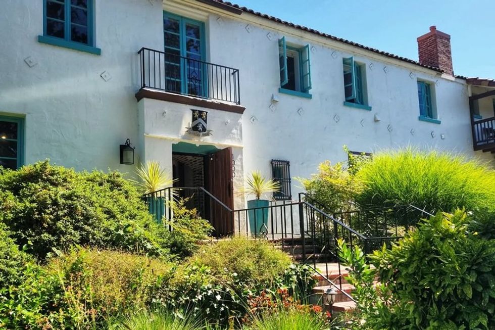 White two-story house with turquoise windows, surrounded by lush greenery under a sunny sky.