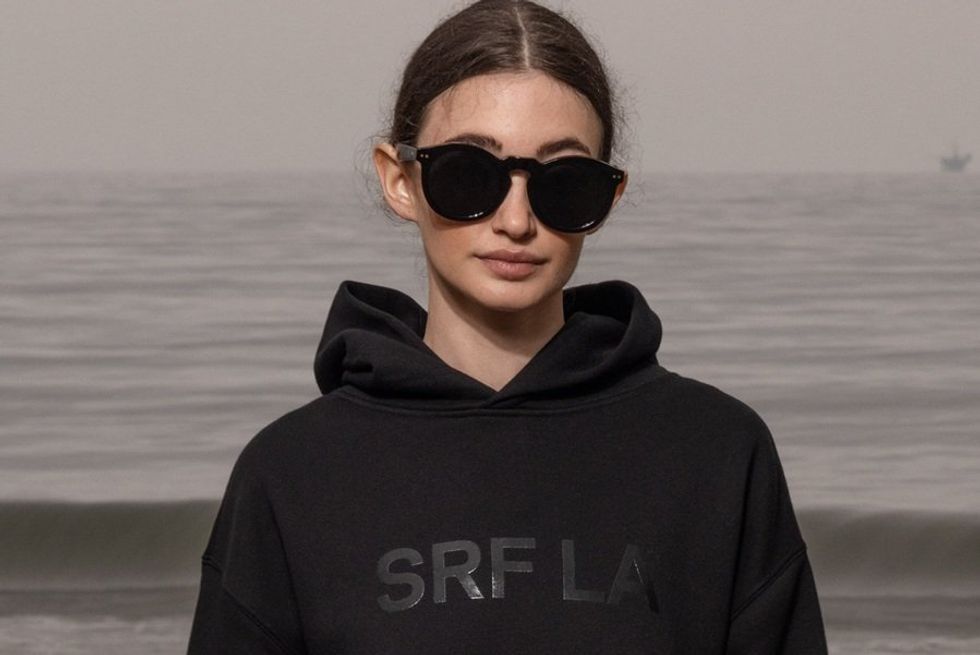 Woman in sunglasses and hoodie standing by the sea.
