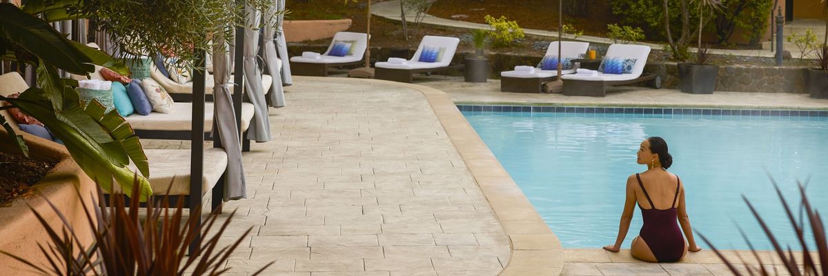Woman in swimsuit sitting by a tranquil pool surrounded by loungers and umbrellas.