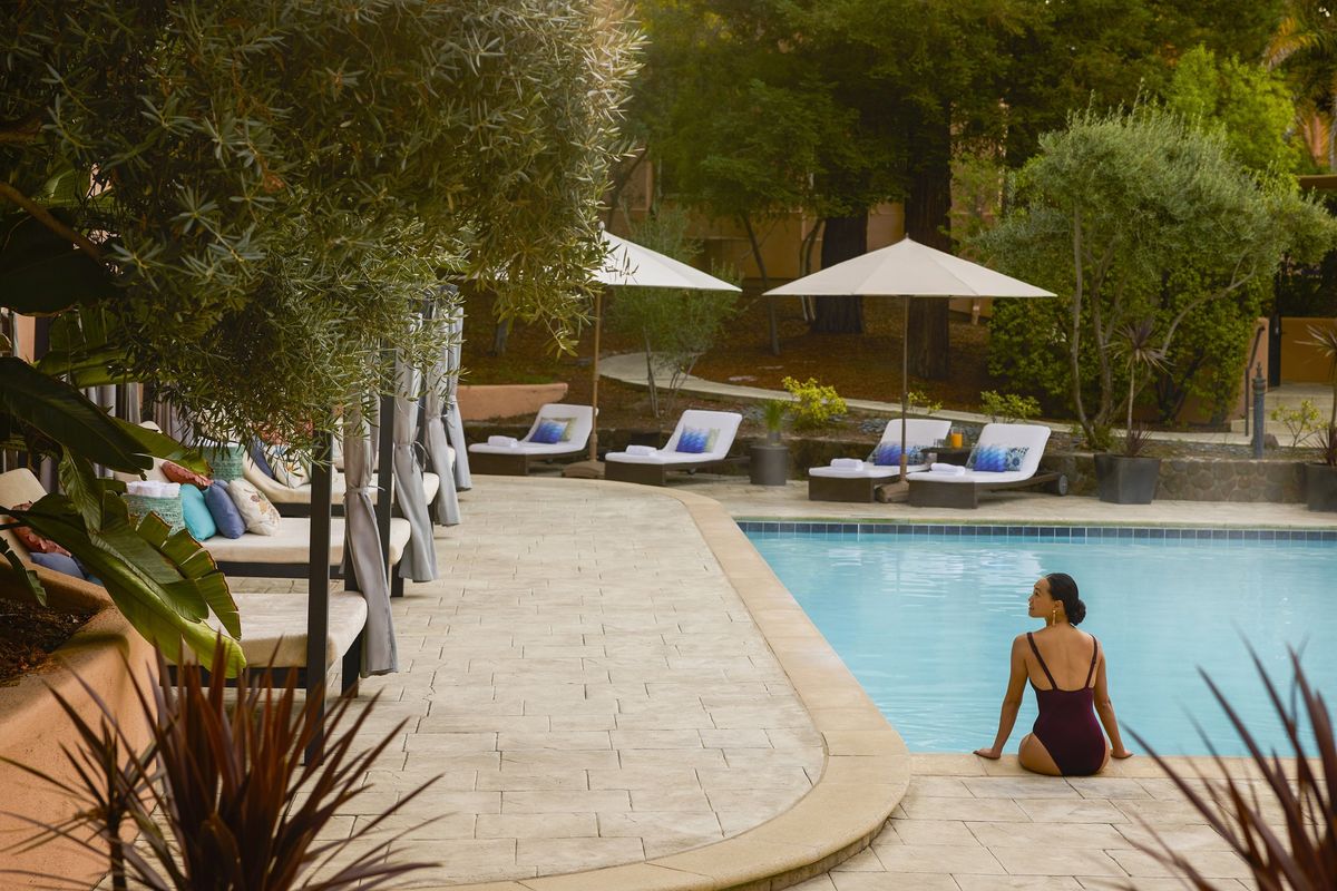 Woman in swimsuit sitting by a tranquil pool surrounded by loungers and umbrellas.