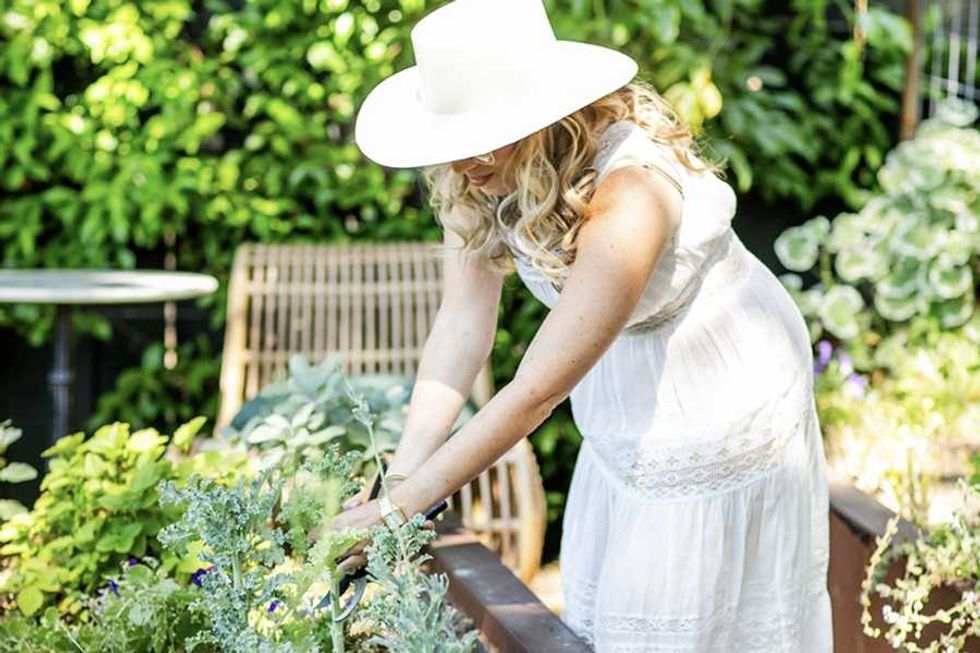 Woman in white dress and hat gardening in a bright, lush garden.