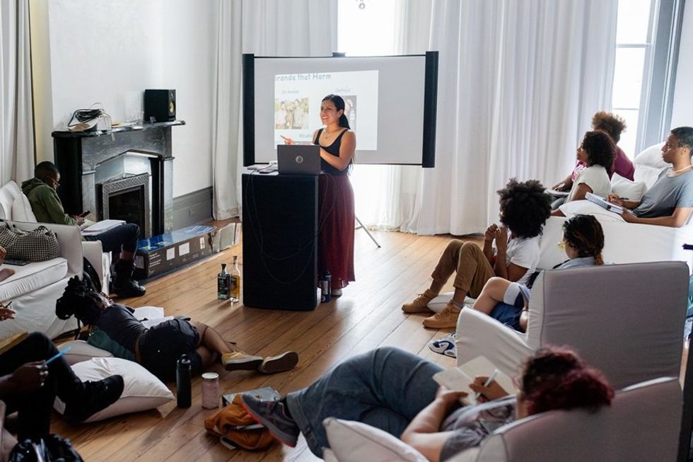Woman leads a presentation in a cozy room with an audience seated on couches and pillows.