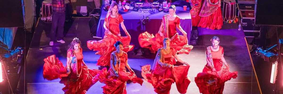 women dancing in red saris on a stage with a dj behind them and a sign that says "Bhangra & Beats Night Market"