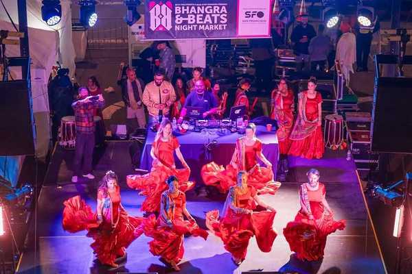 women dancing in red saris on a stage with a dj behind them and a sign that says "Bhangra & Beats Night Market"