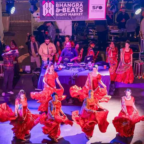 women dancing in red saris on a stage with a dj behind them and a sign that says "Bhangra & Beats Night Market"