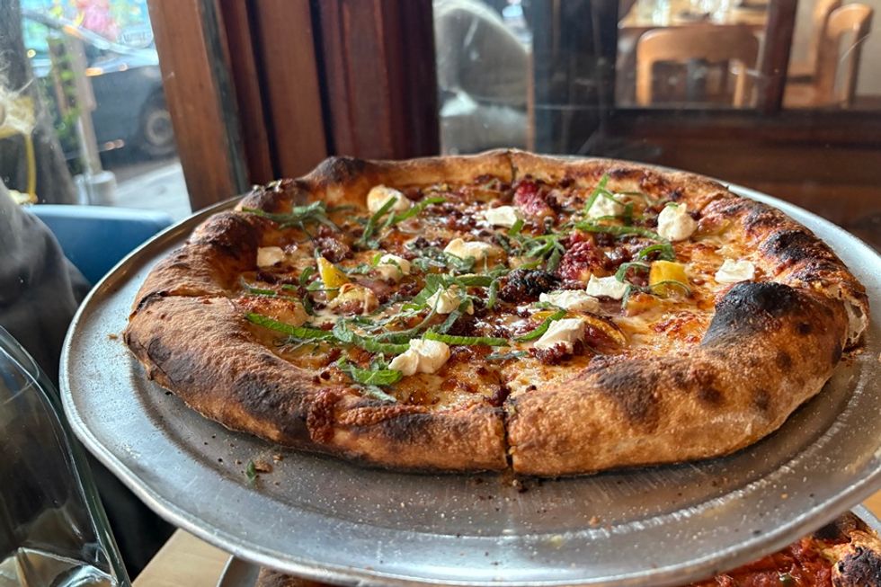 Wood-fired pizza with cheese, herbs, and sun-dried tomatoes on a metal tray.
