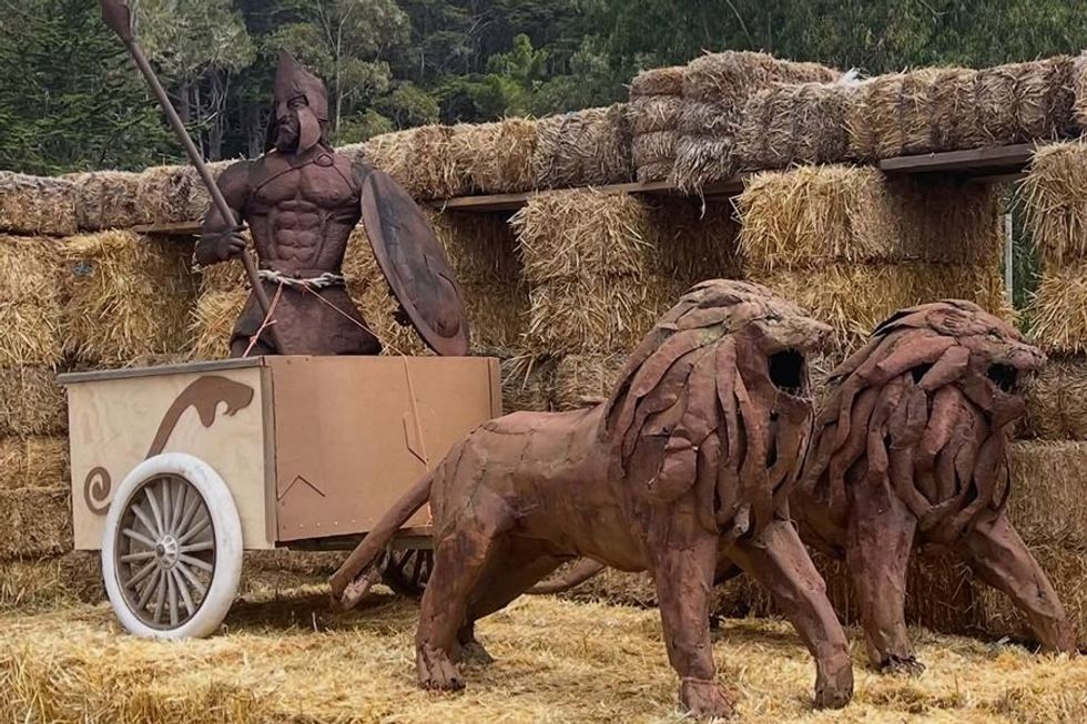 Wooden warrior on a chariot pulled by lion sculptures, surrounded by hay bales.